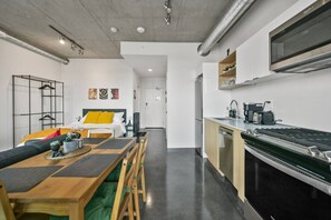 A modern kitchen with a built-in stove, a dining area with a wooden table and chairs, and a bed with a yellow pillow. - Bright Studio | Mins to Saddledome | Skyline Views (Calgary)