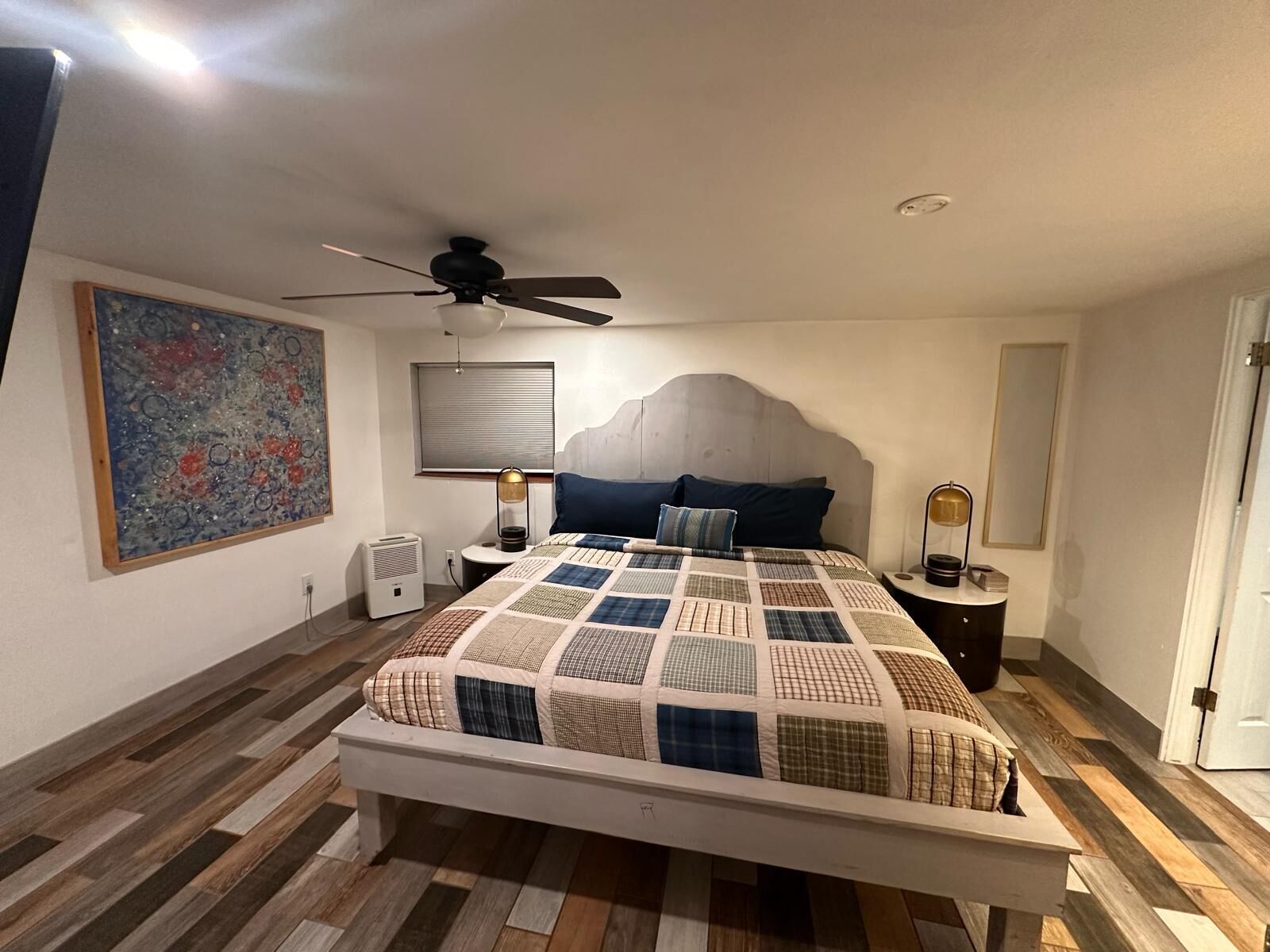 Une chambre à coucher avec un lit, un ventilateur de plafond, un tableau, un radiateur et une fenêtre avec des stores.