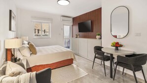 A modern hotel room with a bed, bedside table, lamp, and a dining area with chairs and a table.