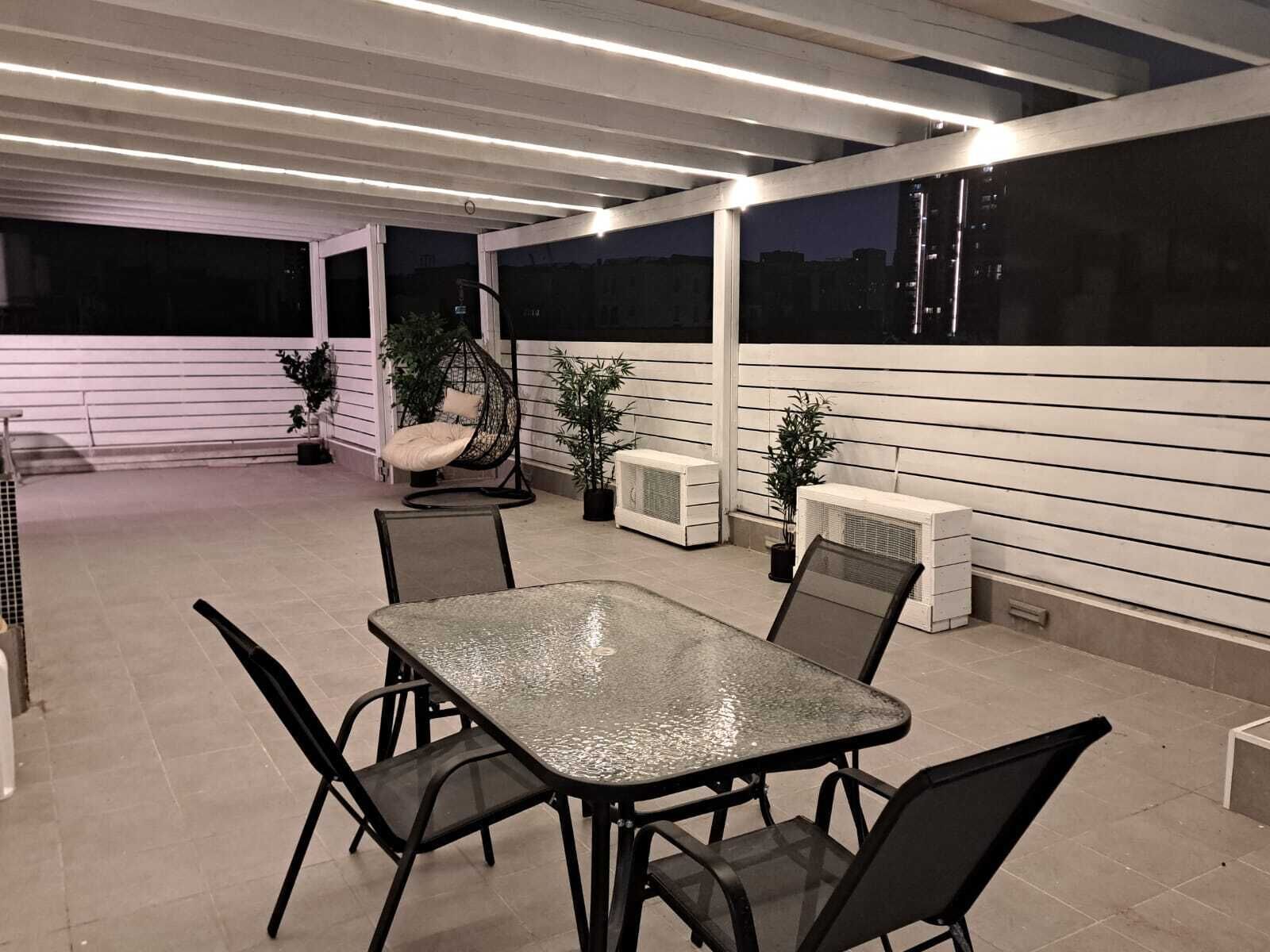En overdækket terrasse med et glasbord og stole, omgivet af krukker med planter og en hængestol.