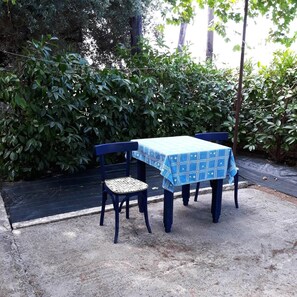 Mesa e duas cadeiras dispostas ao ar livre, com toalha de mesa azul, em meio a vegetação densa.