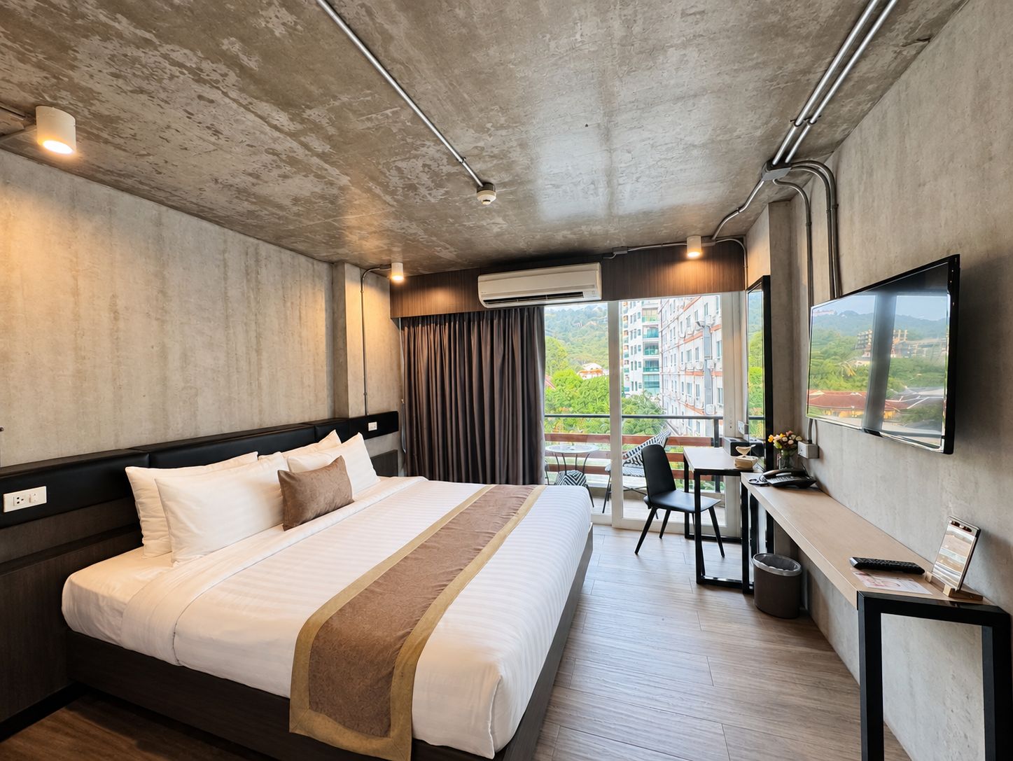 A hotel room with a large bed, a desk with a computer, a TV, and a balcony with a view of buildings.