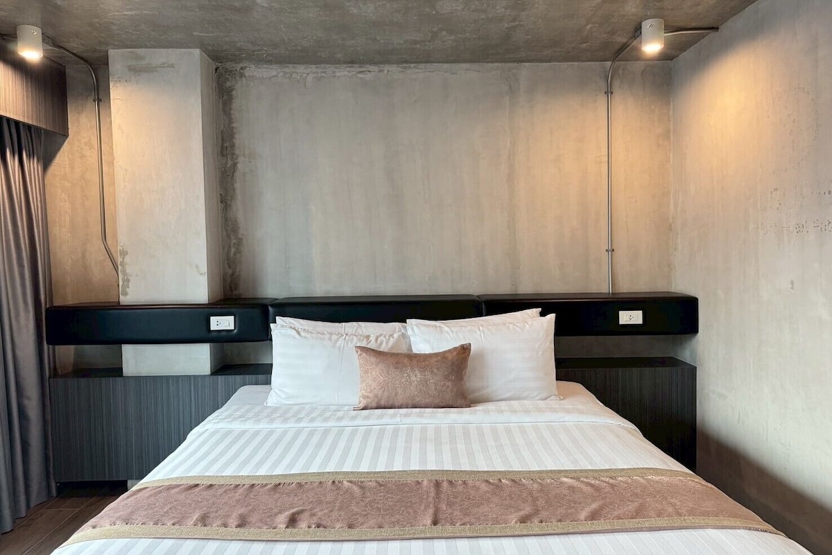 Una habitación de hotel moderna con una cama grande, un cabecero con iluminación integrada y una pared de acento de concreto.