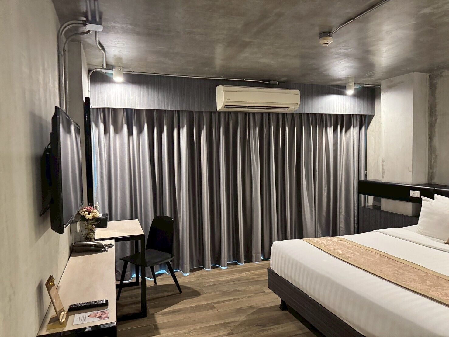 A hotel room with a bed, a desk with a chair, a TV, and an air conditioning unit.