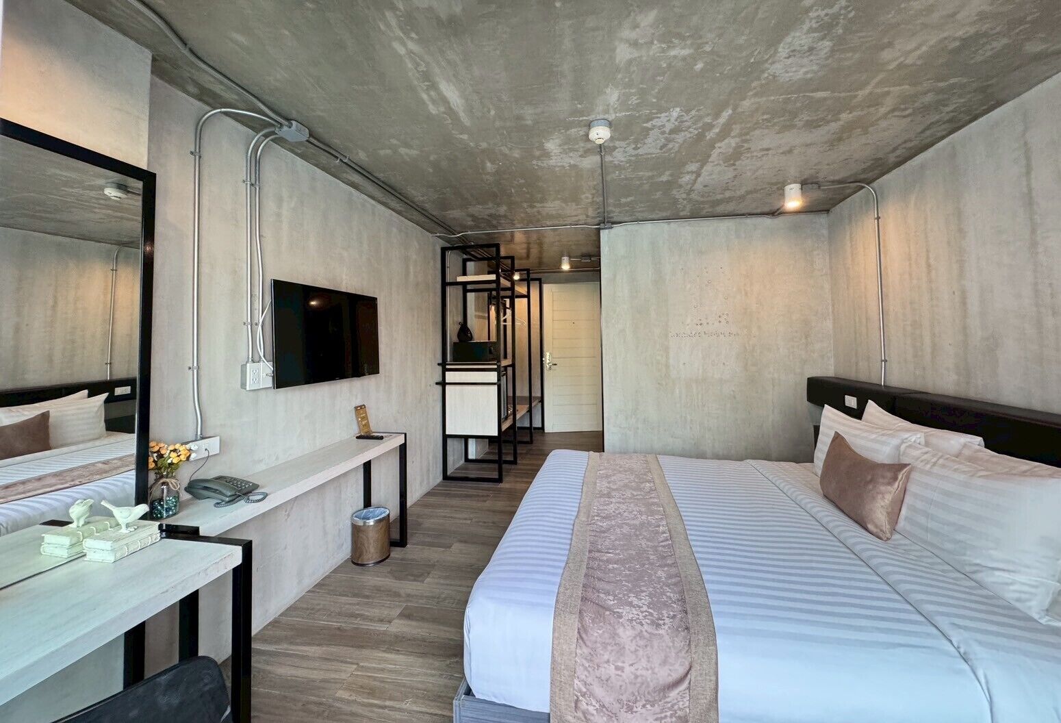 A modern hotel room with a large bed, a desk with a phone and a vase, a TV mounted on the wall, and a bathroom visible through a glass door.