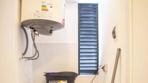 A compact laundry area with a front-loading washing machine, a stacked washer and dryer, a water heater, and a window with blinds. - w* | Modern 1BR in San Isidro (Lima)