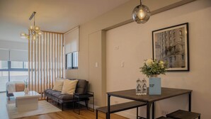 A modern living room with a sofa, coffee table, and a dining table with benches. There is a chandelier, a framed picture on the wall, and a vase with flowers on the dining table. - w* | Modern 1BR in San Isidro (Lima)