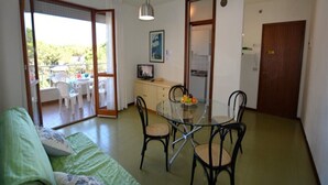 Una sala de estar con una mesa de comedor de vidrio, sillas negras, un sofá verde y un balcón con sillas blancas.
