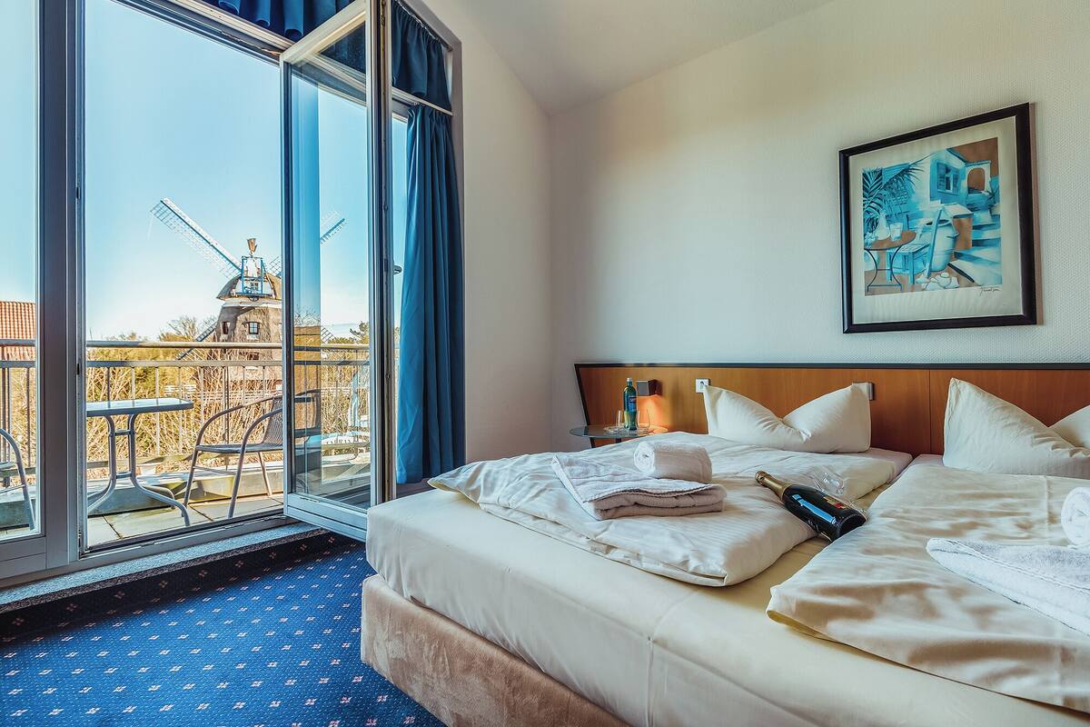 Ein Hotelzimmer mit großem Fenster, einem Bett mit weißen Leinen, einem Gemälde an der Wand und Blick auf eine Windmühle.