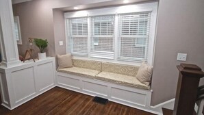 Une pièce avec un banc intégré le long d’une fenêtre dotée de volets blancs, une plante en pot et un plancher en bois.