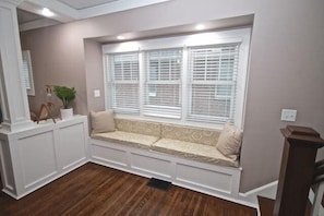 A room with a built-in bench along a window with white shutters, a potted plant, and a wooden floor. - Adorable Broad Ripple Duplex! (Indianapolis)