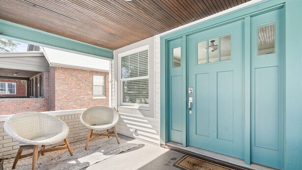 Une porte turquoise avec une fenêtre, une véranda avec deux fauteuils en osier et, au fond, un mur de briques.