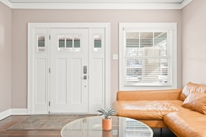 A living room with a beige wall, a white door with glass panels, a window with blinds, and a brown leather sofa.