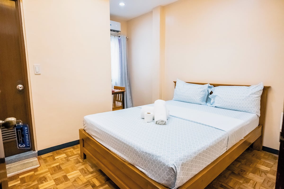 A neatly made bed with a white bedspread, two pillows, and a rolled towel. There is a wooden floor and a door with a number sign.