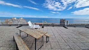 A rooftop terrace with a wooden table and benches, overlooking a coastal area with a clear sky and distant buildings.