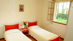 A room with two single beds, a small bedside table with a lamp, and a window with a view of greenery.