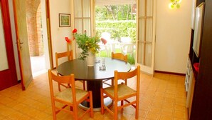 A dining area with a round table, four wooden chairs, a vase with flowers, and a view of the outdoors through a large window.