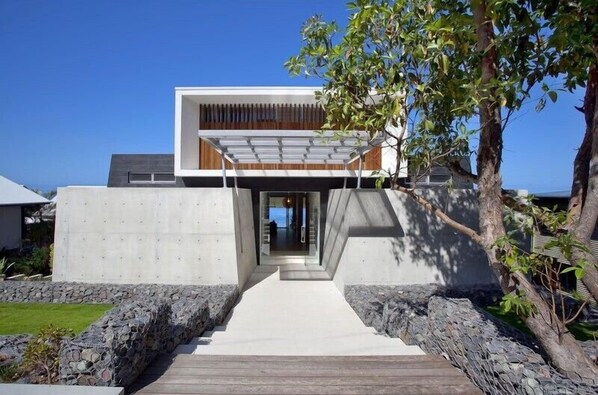 Una casa moderna con fachada de hormigón, terraza de madera y muro de contención de piedra.