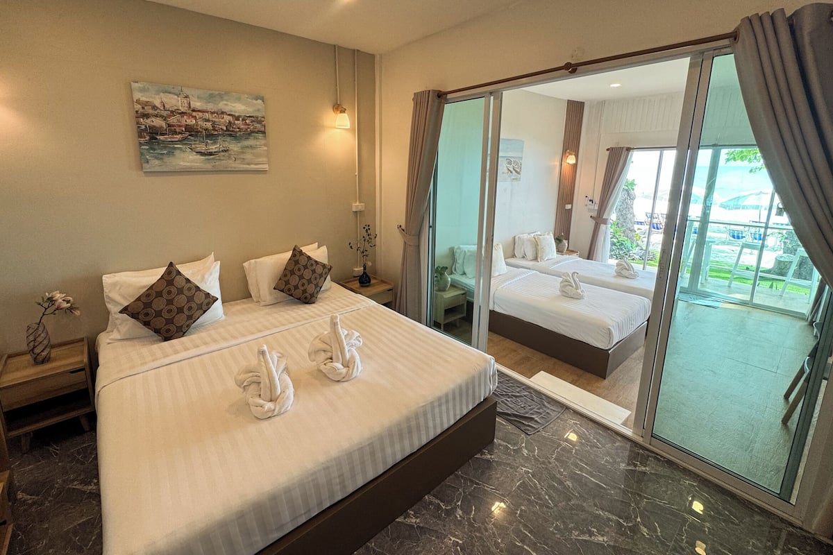 A hotel room with a large bed, two smaller beds, a painting, a vase with flowers, and a view of the outdoors.