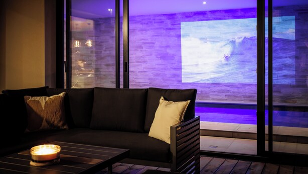 Una sala de estar moderna con un sofá oscuro, una mesita con una vela y un ventanal con vistas a una playa y al océano.