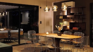 Un comedor moderno con una mesa de madera, sillas de malla metálica, luces colgantes y un puerta de cristal que da al exterior.