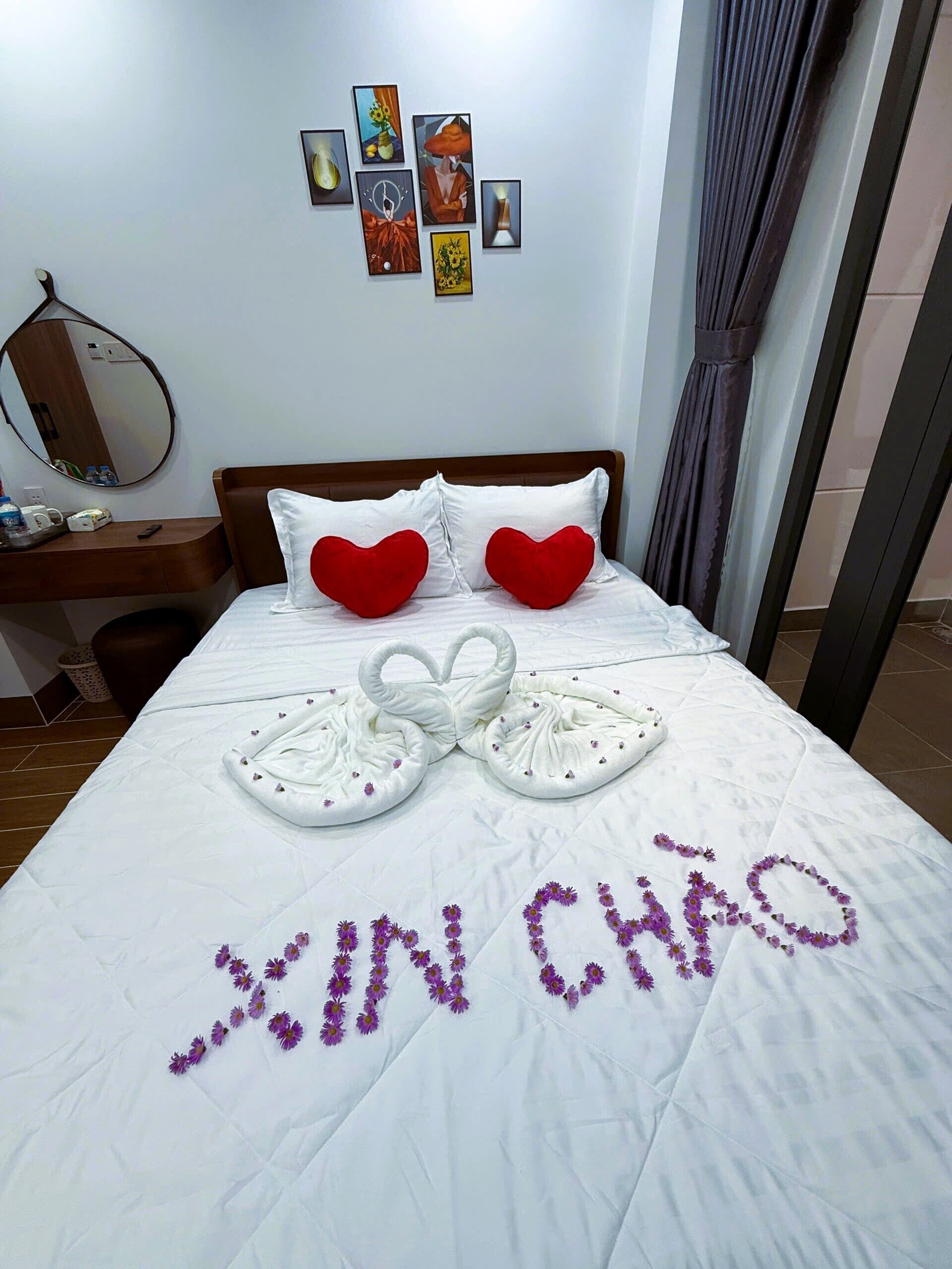 Een hotelkamer met een net opgemaakt bed, hartvormige kussens en een handdoekkunstwerk waarop 'XIN CHAO' staat.