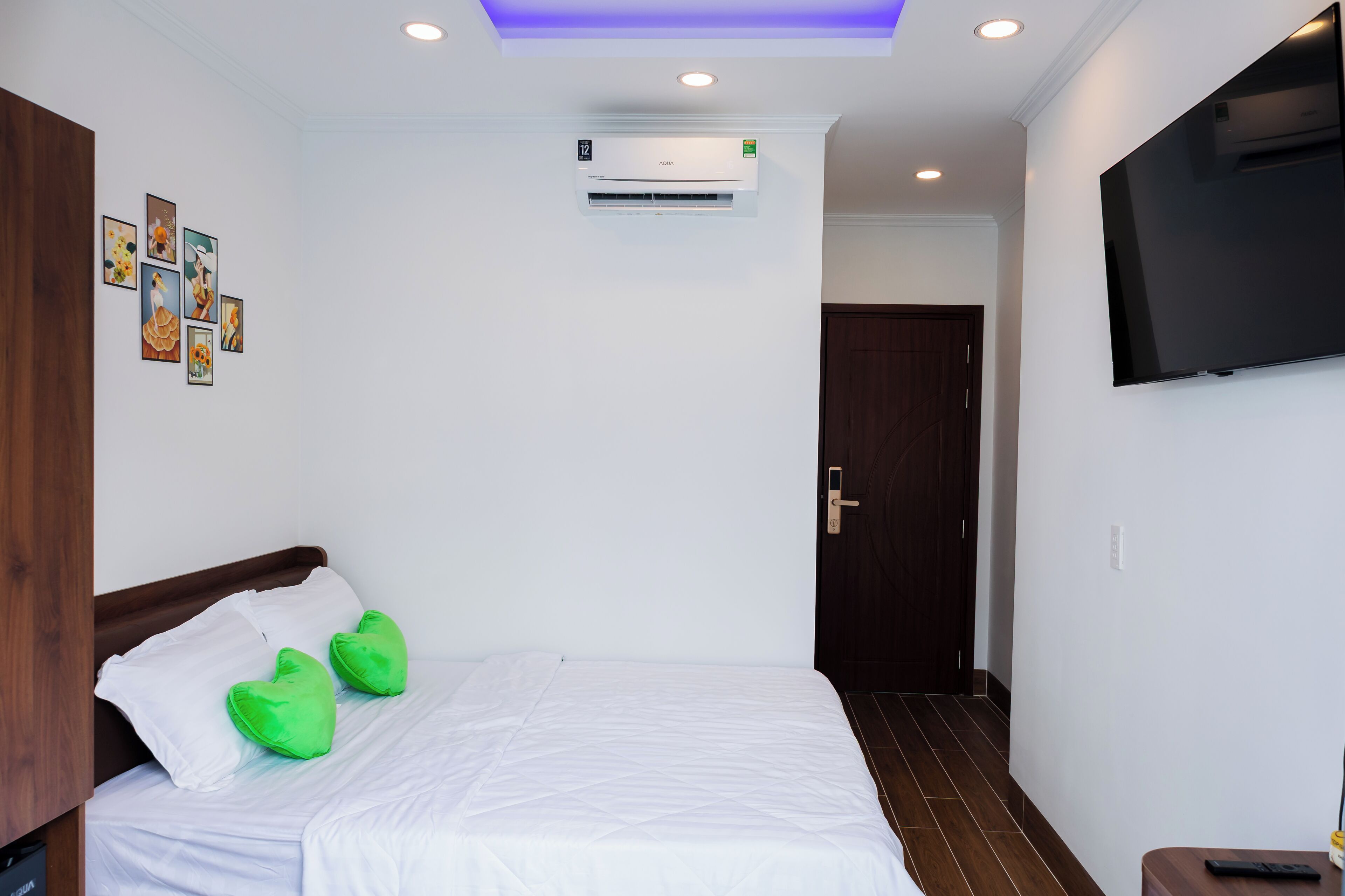 A hotel room with a bed, green heart-shaped pillows, a flat-screen TV, an air conditioning unit, and a door.