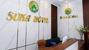 A reception area with a marble wall, two large golden 'Suka Hotel' signs, a wooden reception desk, a brown sofa, and a vase with flowers. - Suka Hotel (Tan Ninh)