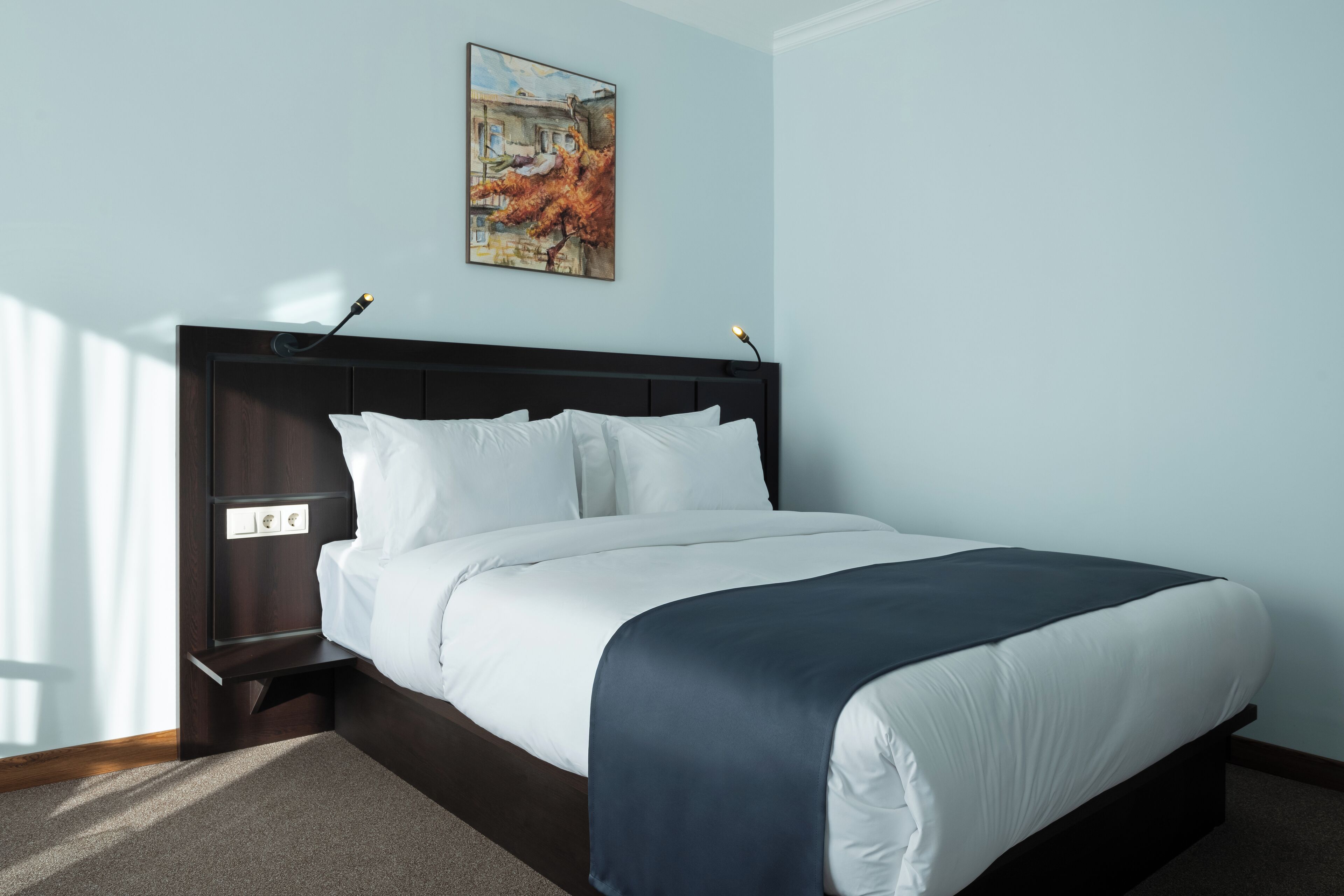 A neatly made bed with white linens and a dark blue throw blanket, a dark wooden headboard, and a painting on the wall.