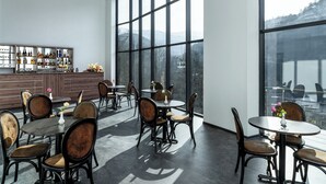 Un área de bar moderna con varias mesas y sillas, ventanas amplias que ofrecen vistas al exterior, y una barra repleta de diferentes botellas.