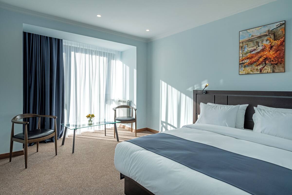 A hotel room with a large bed, a chair, a glass table, and a painting on the wall.