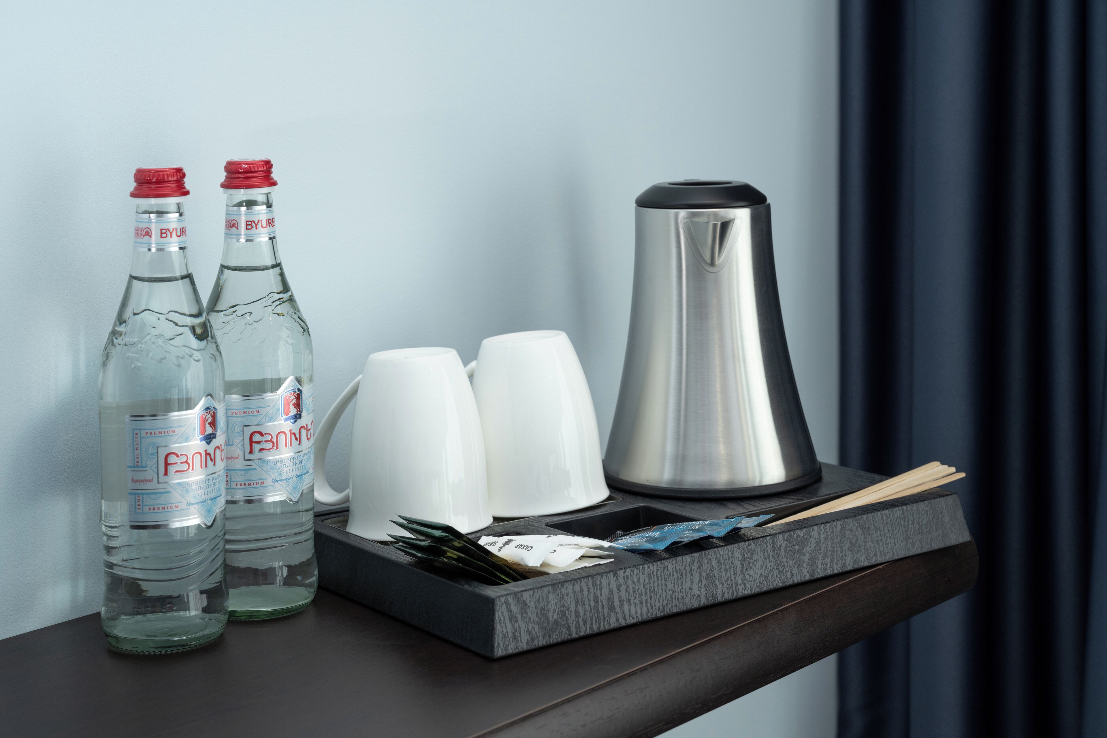 Dos botellas de agua transparente, dos tazas blancas y una cafetera de acero inoxidable sobre una bandeja de madera oscura.