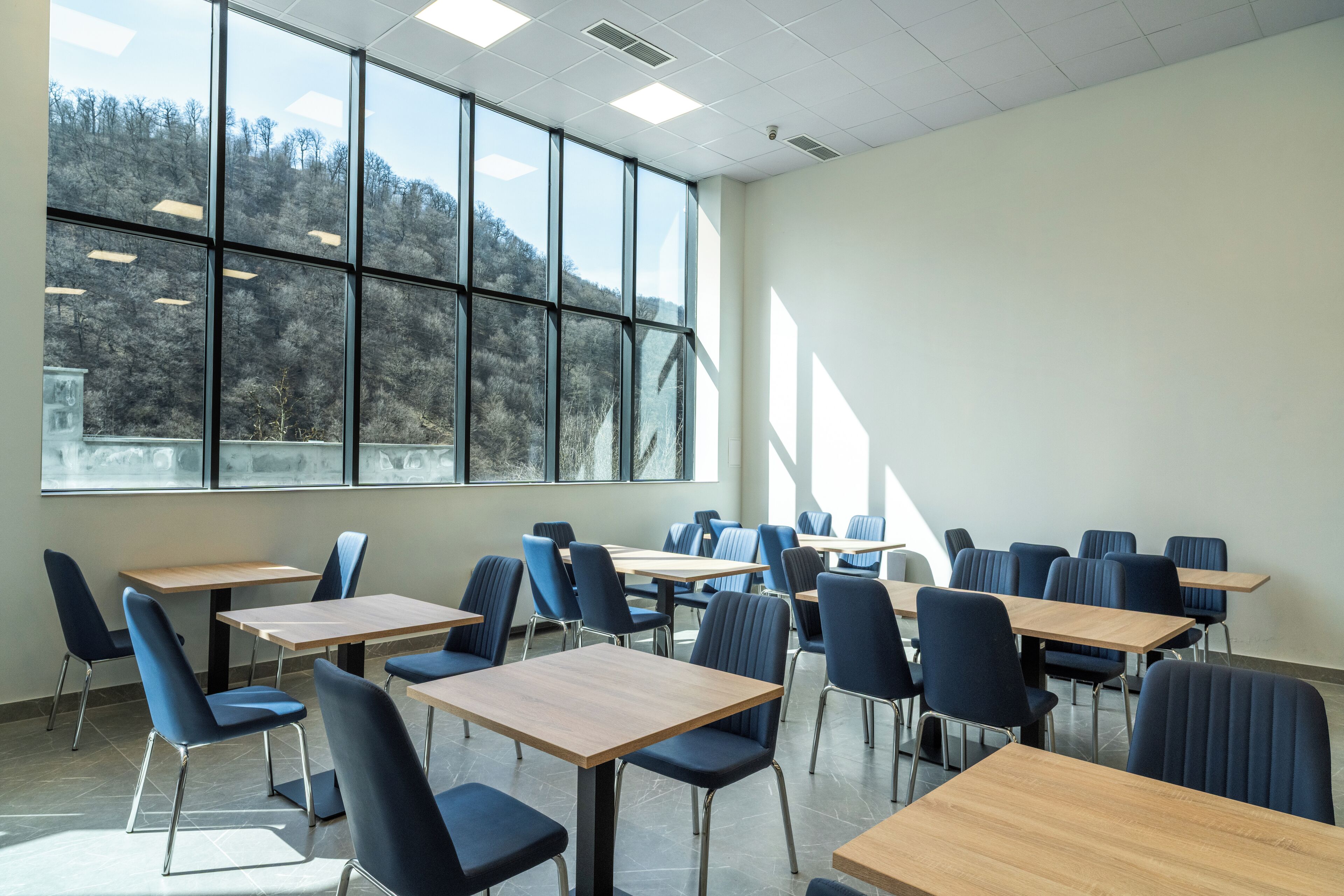 A classroom with multiple desks and chairs, large windows, and a view of trees outside.