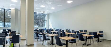 A spacious room with multiple tables and chairs, large windows, and a ceiling with recessed lighting.