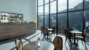 Un bar con mostrador de madera y estanterías llenas de botellas de licor, amplias ventanas con vista a la montaña y mesas redondas con sillas.