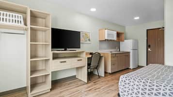 A compact room with a bed, a desk with a chair, a kitchenette, and a wardrobe with open shelves.