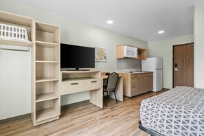 Een kleine kamer met een bed, een bureau met stoel, een kitchenette en een garderobe met open planken.