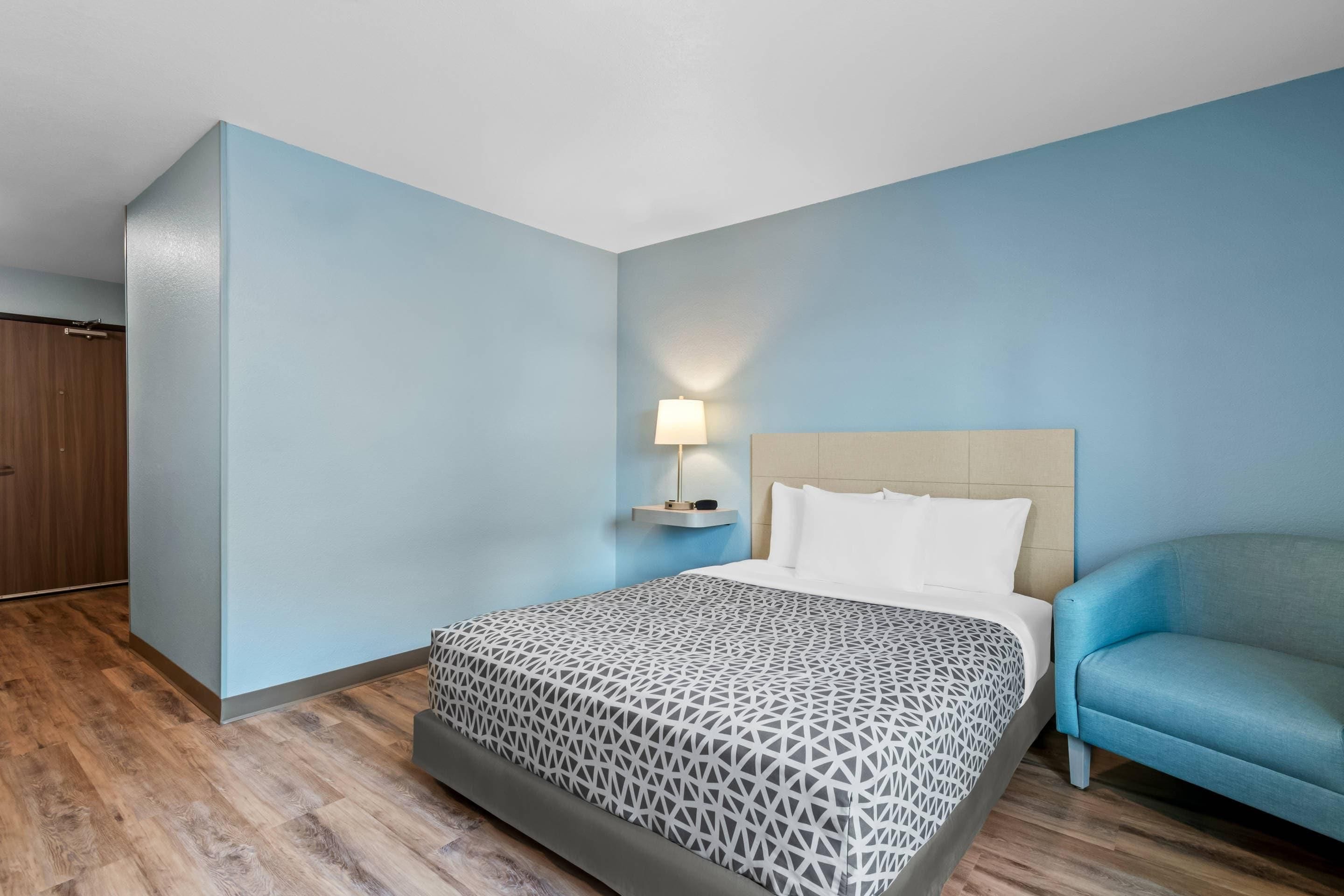 Una habitación de hotel con una cama, una mesita de noche con lámpara, un sillón azul y un armario de madera.