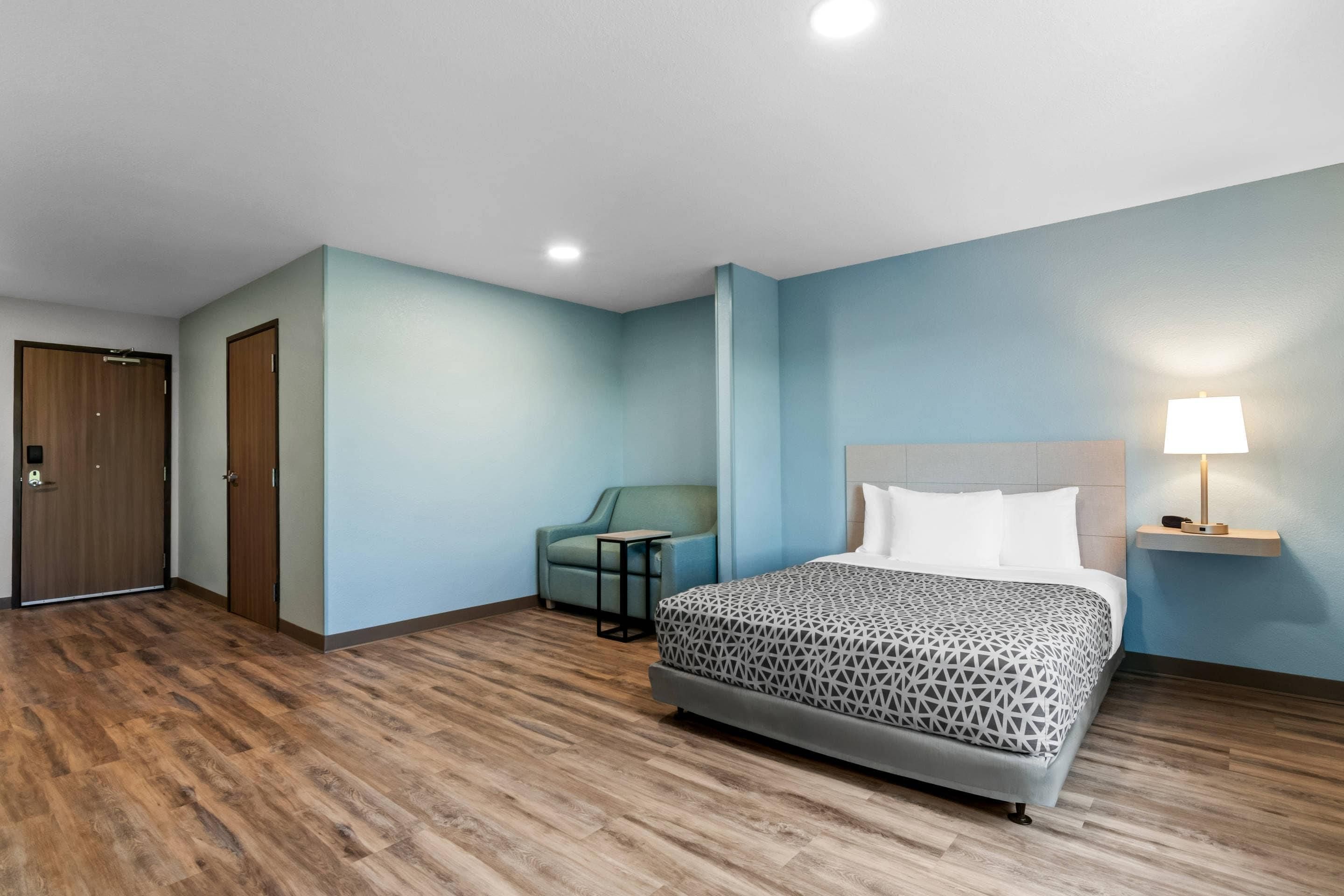 Una habitación de hotel con una cama, una silla, una mesita, una lámpara y una puerta.