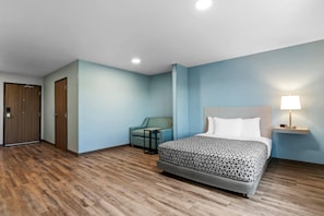 A hotel room with a bed, a chair, a small table, a lamp, and a door.
