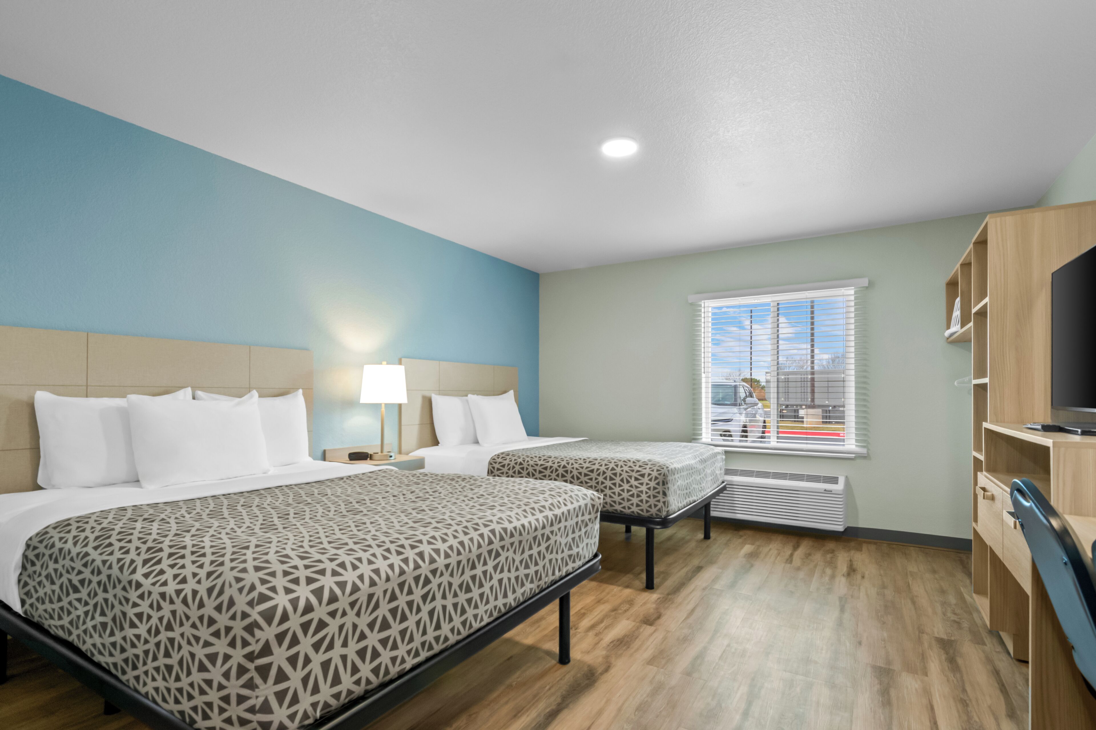 Quarto de hotel com duas camas, televisão, janela com persianas e chão de madeira.