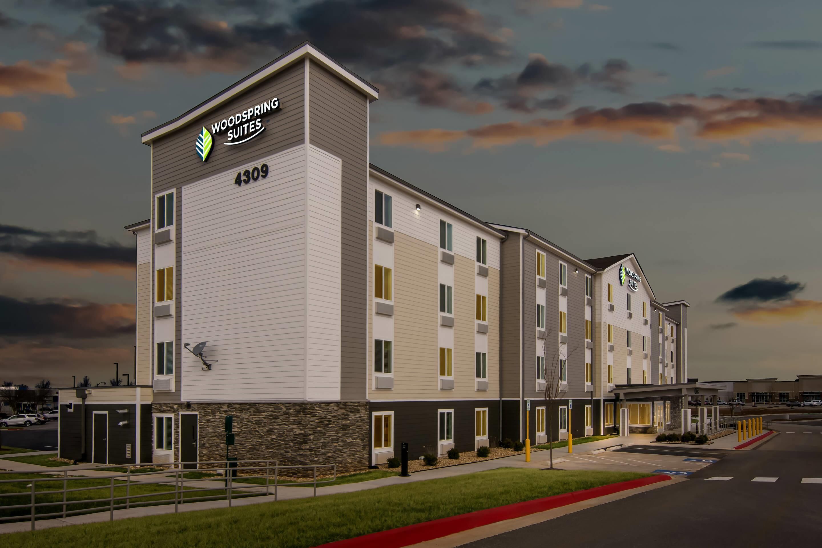 A multi-story hotel with a flat roof, a sign reading 'Woodspring Suites 4309,' and a parking lot in front.