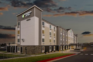 A multi-story hotel with a flat roof, a sign reading 'Woodspring Suites 4309,' and a parking lot in front. - Woodspring Suites Rogers Bentonville (Lowell)