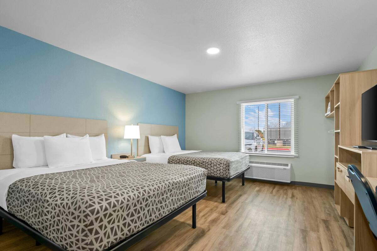 Quarto de hotel com duas camas, uma televisão, janela com persianas e escrivaninha de madeira.