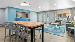 Habitación de hotel moderna con una mesa de comedor de madera, sillas grises, una pared de acento azul, un ventanal con cortinas y una alfombra azul estampada.