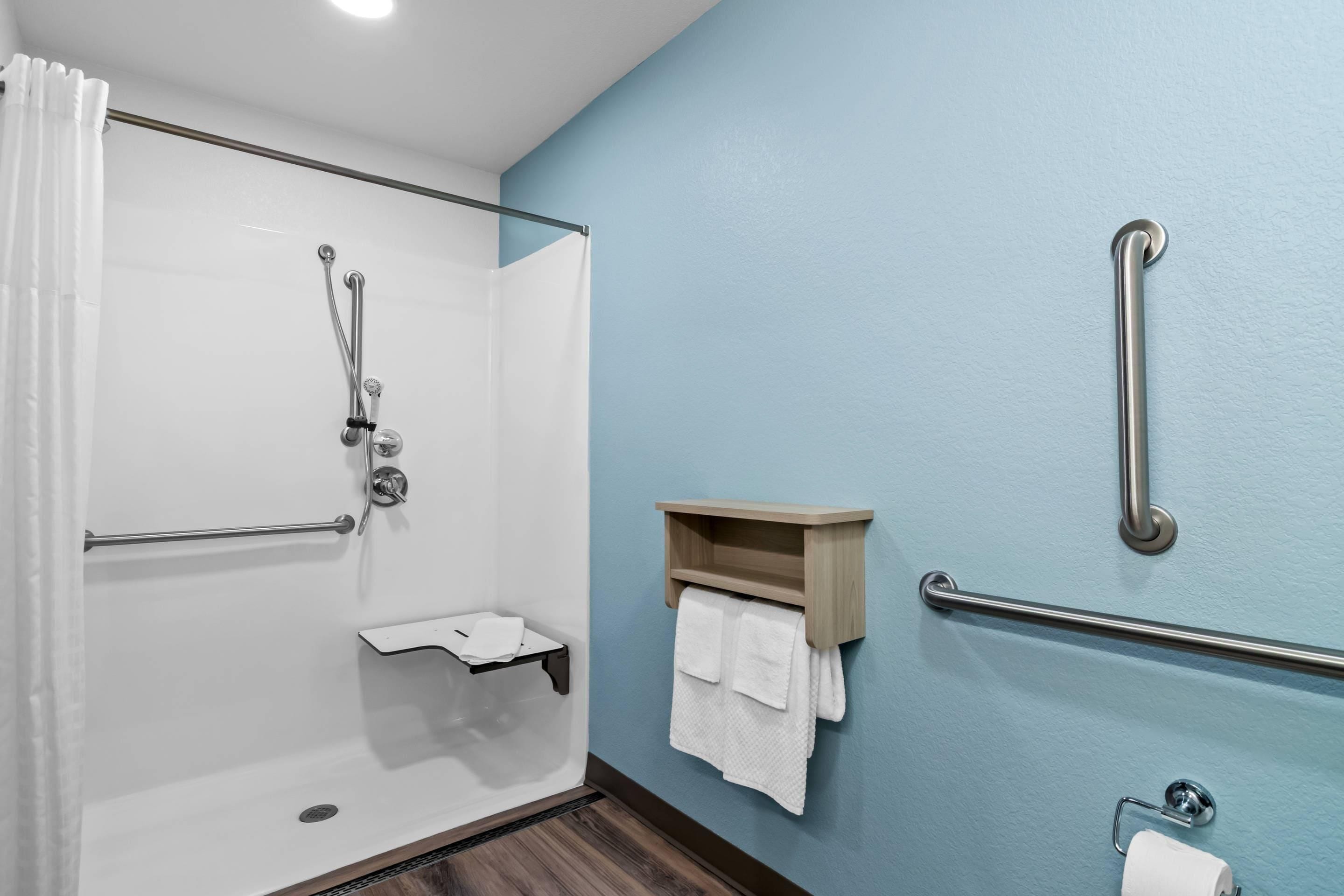 Ducha con puerta de vidrio, estante de madera con toallas, pasamanos y dispensador de papel higiénico.