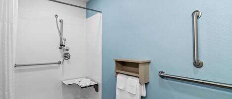 Ducha con puerta de vidrio, estante de madera con toallas, pasamanos y dispensador de papel higiénico.