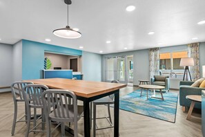 Habitación de hotel moderna con una mesa de comedor de madera, sillas grises, una pared de acento azul, un ventanal con cortinas y una alfombra azul estampada.