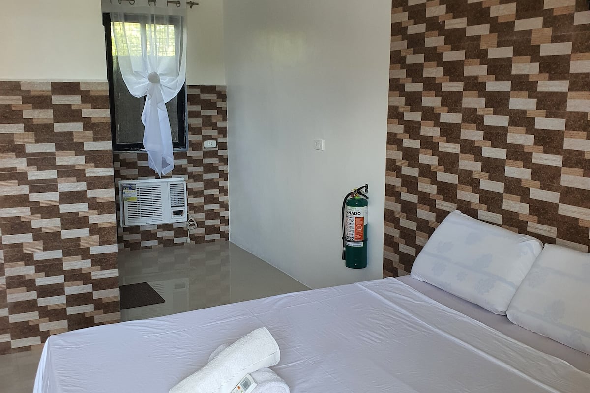 Una camera d'albergo con un letto, un condizionatore d'aria a parete, un estintore e una finestra con tende.