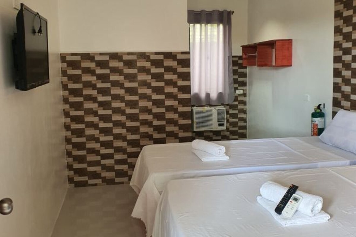 A hotel room with two beds, a wall-mounted TV, a window with curtains, a wall-mounted heater, and a small shelf with toiletries.